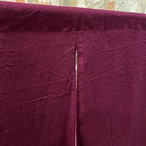 Pode incluir: Uma cortina noren de tecido em tom bord&ocirc; profundo com uma superf&iacute;cie s&oacute;lida e texturizada, apresentando uma fenda vertical ao centro. O tecido possui acabamento fosco e vincos vis&iacute;veis, suspensa por uma haste horizontal na parte superior.