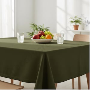Puede incluir: Un mantel cuadrado verde oliva cubre una mesa de madera. Un cuenco blanco con fruta, incluyendo manzanas rojas, uvas verdes y una naranja, est&aacute; en el centro. Dos vasos transparentes con agua est&aacute;n a cada lado del cuenco. Una planta est&aacute; en el fondo.
