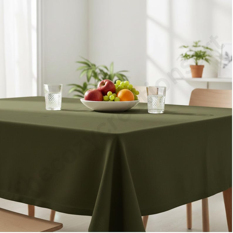Puede incluir: Un mantel cuadrado verde oliva cubre una mesa de madera. Un cuenco blanco con fruta, incluyendo manzanas rojas, uvas verdes y una naranja, est&aacute; en el centro. Dos vasos transparentes con agua est&aacute;n a cada lado del cuenco. Una planta est&aacute; en el fondo.