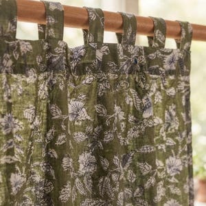 Könnte beinhalten: Ein olivgrüner Vorhang mit einem sich wiederholenden weißen Blumen- und Blattmuster. Der Vorhang ist an einer Holzstange durch eine Reihe von Stoffschlaufen an der Oberkante befestigt, was einen gerafften, gefalteten Effekt auf der Stoffoberfläche erzeugt.