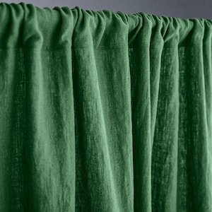 May include: Close-up of a green linen curtain. The fabric is textured and has vertical folds. The curtain is hanging from a rod, with the top gathered. The color is a deep, rich green.