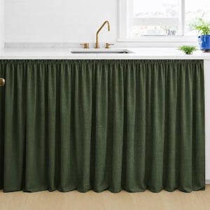 Pode incluir: Uma saia de pia de cozinha verde escuro feita de tecido texturizado. A saia é franzida na parte superior e pende em dobras verticais. Uma torneira dourada e uma bancada branca são visíveis acima.