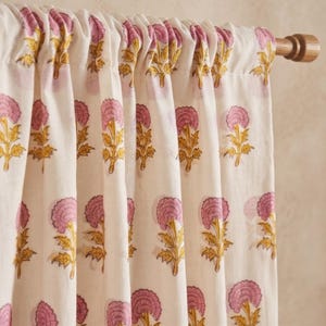 May include: A cream-coloured curtain featuring a repeating floral pattern of pink blossoms with yellow stems and leaves. The fabric is gathered along a wooden curtain rod, creating vertical folds that display the consistent botanical print.