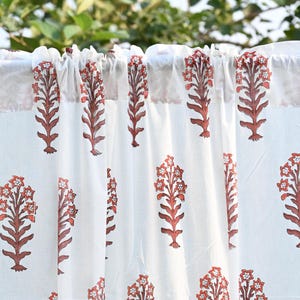 Puede incluir: Una cortina blanca con un patrón repetitivo estampado en bloque de motivos florales de color terracota. La tela está fruncida en la parte superior a lo largo de una barra, creando pliegues verticales que muestran el diseño botánico simétrico.