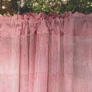 May include: Red and white striped curtain panel. The fabric is gathered at the top, creating a ruffled effect. The vertical stripes are evenly spaced, and the curtain appears to be made of a lightweight material. The background is blurred, suggesting an outdoor setting.