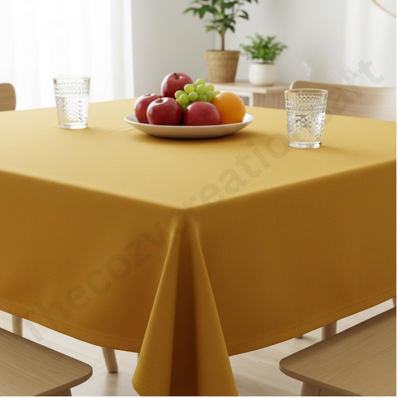 Puede incluir: Un mantel cuadrado de color amarillo mostaza cubre una mesa de madera. Un plato blanco con fruta, que incluye manzanas rojas, uvas verdes y una naranja, est&aacute; en el centro. Dos vasos transparentes est&aacute;n sobre la mesa.