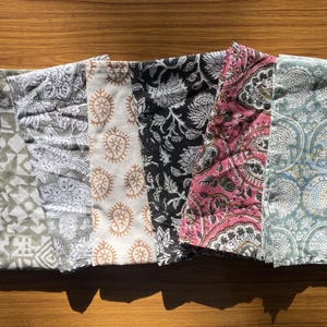 May include: A set of six rectangular fabric napkins featuring diverse block-printed patterns. The designs include olive green geometric shapes, grey floral motifs, tan paisley, black floral, pink paisley, and blue circular patterns on white fabric.