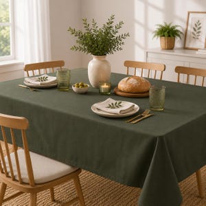 Puede incluir: Un mantel rectangular de color verde oliva oscuro sólido sobre una mesa de comedor. La mesa está puesta con platos blancos, servilletas dobladas, vasos verdes texturizados, una hogaza de pan redonda en una tabla de madera, un cuenco y un jarrón.