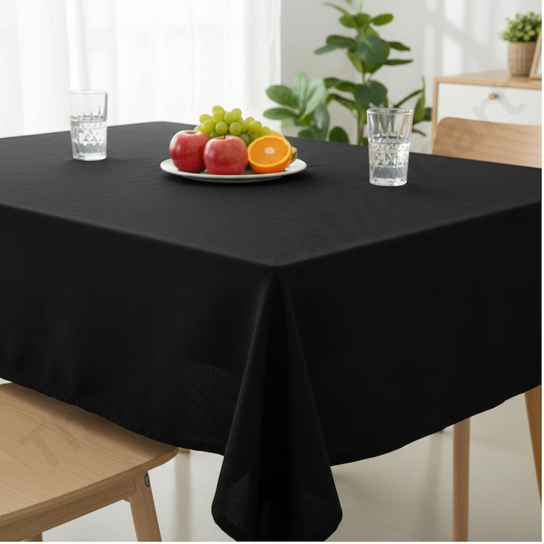 Puede incluir: Un mantel negro rectangular cubre una mesa de madera. Se ven dos vasos de agua y un plato de fruta, que incluye manzanas rojas, una rodaja de naranja y uvas verdes. La escena tiene buena iluminaci&oacute;n.