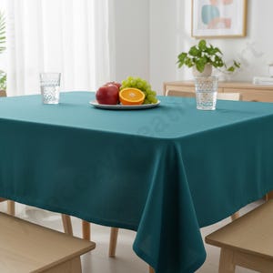 Puede incluir: Un mantel rectangular de color verde azulado cubre una mesa de madera. Un plato blanco contiene una manzana roja, una rodaja de naranja y uvas verdes. Dos vasos transparentes y una peque&ntilde;a planta tambi&eacute;n est&aacute;n sobre la mesa. El mantel es de tela lisa.
