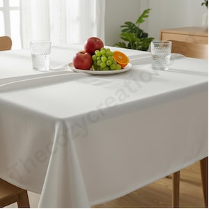 Puede incluir: Un mantel blanco cubre una mesa de madera, con un plato de manzanas rojas, uvas verdes y una rodaja de naranja. Dos vasos transparentes con dise&ntilde;os texturizados est&aacute;n colocados sobre la mesa. La escena sugiere un comedor o una cocina.