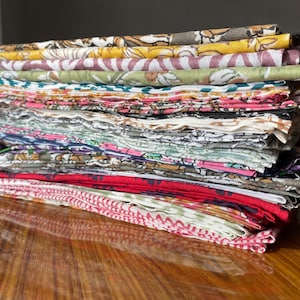 May include: A tall stack of folded rectangular fabric pieces featuring a variety of colorful floral and geometric block-printed patterns in shades of yellow, green, pink, red, blue, and black, layered neatly to display the diverse textile designs.