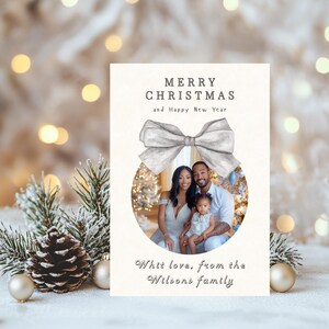 Könnte beinhalten: Eine weiße Weihnachtskarte mit einer silbernen Schleife und einem Foto einer dreiköpfigen Familie vor einem Weihnachtsbaum. Die Karte lautet "Frohe Weihnachten und ein glückliches neues Jahr" und "Mit Liebe, von der Familie Wilson".