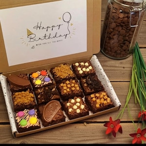 May include: A selection of decorated brownies in a box, with toppings like chocolate and sweets. The box has a card that reads "Happy Birthday". A jar of chocolate chips and red flowers are also visible.