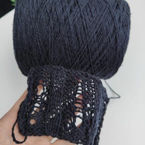 May include: A large, cylindrical ball of dark navy blue yarn positioned above a rectangular swatch of the same yarn. The swatch features a repeating openwork lace pattern with vertical columns of eyelets and knit stitches in a dark navy blue color.