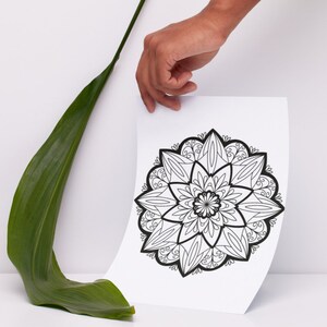 May include: A black and white mandala design printed on a white sheet of paper. The paper is being held by a hand.