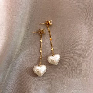 Könnte beinhalten: Ein Paar goldene Ohrringe mit einem kleinen, klaren Kristall oben und einem weißen herzförmigen Anhänger unten.