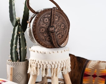May include: A brown leather handbag with a round, tooled design. The bag is sitting on a white stool with fringe. The stool is on a brown and white cowhide rug.