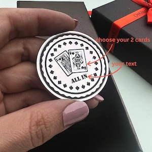 Op de afbeelding: Een zilverkleurige pokerchip met een speelkaartenontwerp. De chip heeft twee kaarten en de woorden "ALL IN". De chip wordt in een hand gehouden, met een zwarte geschenkdoos op de achtergrond.