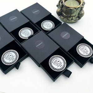 Può includere: Diverse scatole regalo nere, ognuna contenente un gettone da poker argentato. I gettoni presentano un disegno di mano di carte e la scritta "Groomsman 2025". Le scatole hanno la scritta "CHARMAYS".