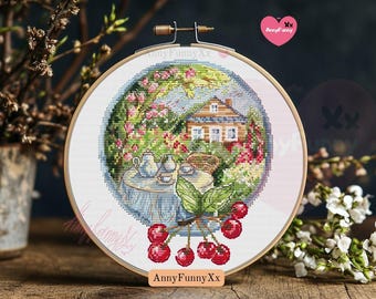May include: A round embroidery hoop with a cross-stitch design of a house with a garden, a table with teacups, and a cherry tree. The design is in shades of blue, green, pink, and white. The hoop is on a wooden surface.