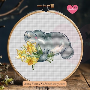 May include: Cross stitch pattern featuring a grey manatee holding a bouquet of yellow daffodils. The pattern is on white Aida cloth in a wooden embroidery hoop.
