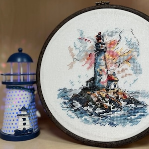 May include: A white embroidery hoop with a cross-stitch design of a lighthouse on a rocky shore with blue and white waves. The sky is a light blue with pink and orange clouds.