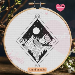 May include: Black and white cross stitch pattern of a mountain landscape with a full moon and stars in a diamond shape. The pattern is on a white Aida cloth in a wooden embroidery hoop.