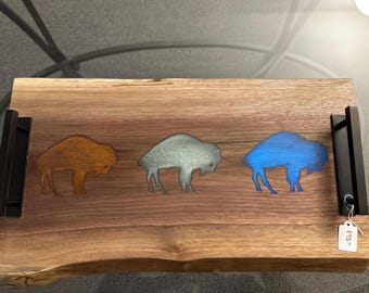 May include: A wooden serving tray with three bison silhouettes in orange, gray, and blue. The tray has black handles and a natural wood grain finish. A price tag hangs from one of the handles.