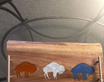 May include: A wooden serving tray with three metal bison silhouettes in rust, silver, and blue. The tray has silver handles and a natural wood grain finish. The tray is rectangular.