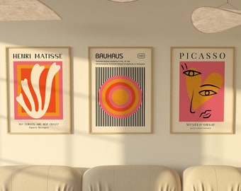 May include: Three framed art prints with minimalist designs in pink, orange, and yellow. The prints are titled "Henri Matisse", "Bauhaus", and "Picasso" and are hung on a white wall above a beige sofa.
