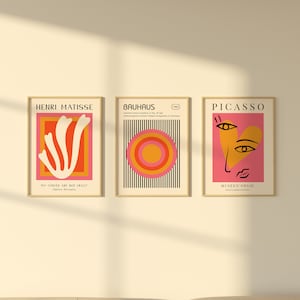 K&ouml;nnte beinhalten: Drei gerahmte Kunstdrucke mit minimalistischen Designs. Der linke Druck zeigt eine wei&szlig;e abstrakte Form auf einem roten und orangefarbenen Hintergrund mit dem Text "Henri Matisse" und "Meine Kurven sind nicht verr&uuml;ckt." Der mittlere Druck zeigt einen schwarz-wei&szlig; gestreiften Hintergrund mit einem roten, orangefarbenen und gelben Kreis in der Mitte. Der Text "Bauhaus" und "1923" befindet sich oben auf dem Druck. Der rechte Druck zeigt einen rosa Hintergrund mit einer gelben Herzform, die ein stilisiertes Gesicht mit schwarzen Augen und einer schwarzen Nase enth&auml;lt. Der Text "Picasso" und "Mus&eacute;e d'Orsay" befindet sich oben auf dem Druck.