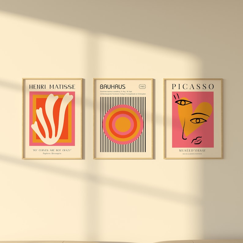 K&ouml;nnte beinhalten: Drei gerahmte Kunstdrucke mit minimalistischen Designs. Der linke Druck zeigt eine wei&szlig;e abstrakte Form auf einem roten und orangefarbenen Hintergrund mit dem Text "Henri Matisse" und "Meine Kurven sind nicht verr&uuml;ckt." Der mittlere Druck zeigt einen schwarz-wei&szlig; gestreiften Hintergrund mit einem roten, orangefarbenen und gelben Kreis in der Mitte. Der Text "Bauhaus" und "1923" befindet sich oben auf dem Druck. Der rechte Druck zeigt einen rosa Hintergrund mit einer gelben Herzform, die ein stilisiertes Gesicht mit schwarzen Augen und einer schwarzen Nase enth&auml;lt. Der Text "Picasso" und "Mus&eacute;e d'Orsay" befindet sich oben auf dem Druck.