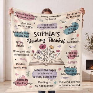 Puede incluir: Una manta de color crema con el texto 'SOPHIA'S Reading Blanket' en el centro sobre una ilustración de un libro abierto con flores coloridas. La manta incluye varias citas y frases relacionadas con los libros en texto negro.