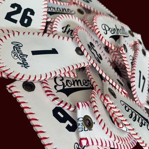 May include: A pile of white leather baseball-themed keychains with red stitching. The keychains feature different numbers, names, and logos, including "Rawlings", "Gomer", and "Penhall".