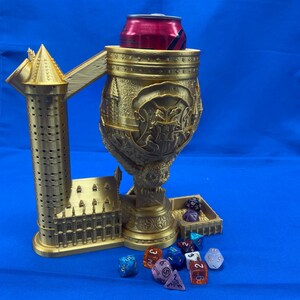 Puede incluir: Un accesorio de juego de mesa impreso en 3D de color dorado con una torre, una copa y una bandeja para dados. La copa tiene una lata dentro y la bandeja para dados tiene varios dados de colores.