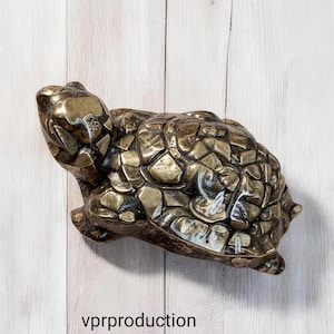 May include: A metallic bronze-toned turtle figurine with a textured, segmented shell and a smooth head. The surface features a weathered, antiqued finish with dark highlights in the crevices. The text vprproduction is visible below the turtle.