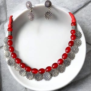 Puede incluir: Un collar de cuentas rojas y un juego de pendientes a juego. El collar presenta cuentas rojas ovaladas y colgantes plateados en forma de flor. Los pendientes tienen un diseño similar, con una parte superior circular plateada y un colgante de flor.