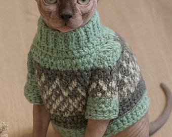 May include: A Sphynx cat wearing a handmade, green, gray, and white patterned sweater. The cat has large ears and green eyes. The sweater has a turtleneck design and covers the cat's torso and arms.