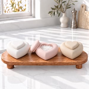 May include: Three heart-shaped containers with lids, colored white, light pink, and beige, arranged on a small, oval, wooden riser with four short legs. The pink container is open, revealing white granular contents inside the hollow center.