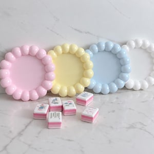 Può includere: Quattro vassoi circolari con un bordo a bolle in colori pastello rosa, giallo, blu e bianco. Davanti sono sparse sei piccole tessere rettangolari da mahjong, ognuna con una parte superiore bianca con illustrazioni colorate e una base rosa e bianca.