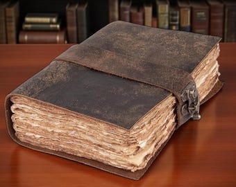May include: A thick, rectangular journal bound in dark brown leather with a distressed texture. The book features a wide leather strap closure secured by a metal latch. The pages are thick, cream-colored, and have uneven, deckled edges.