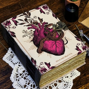 May include: A rectangular journal with a cream-coloured cover featuring a dark pink anatomical heart illustration entwined with a snake, surrounded by dark pink floral motifs. The word INAMORATA is printed below the heart. It has a metal latch.
