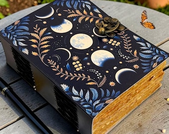 Puede incluir: Un diario azul oscuro con un diseño celestial que presenta fases lunares y elementos botánicos. El diario tiene un cierre decorativo y páginas de aspecto envejecido. Una pluma negra descansa junto al diario sobre una superficie de madera.
