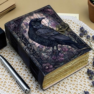 Peut inclure: Un journal rectangulaire avec une couverture sombre représentant un corbeau noir perché dans un cadre de vitrail entouré de fleurs violettes. Le livre possède une fermeture à loquet métallique et des pages épaisses, texturées et crème.