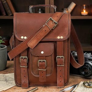 May include: A brown leather satchel with a top handle, shoulder strap, and multiple buckle closures. The bag has a front pocket and brass-colored hardware. A vintage camera and other accessories are visible in the background.