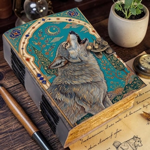 May include: A rectangular journal with a teal cover featuring a detailed illustration of a howling wolf, a crescent moon, and floral patterns. The book has a metallic latch closure and gold-toned page edges, resting on a wooden surface with a pen.