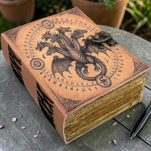 May include: A tan leather journal featuring a dragon illustration and runic symbols. The cover has a detailed black dragon design and decorative borders. A metal clasp secures the journal, and a black pen lies beside it on a wooden table.