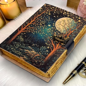 May include: A leather-bound journal with a celestial design featuring a full moon, trees, and stars. The journal has a clasp closure and aged paper. A black and gold pen with a compass charm rests beside it.