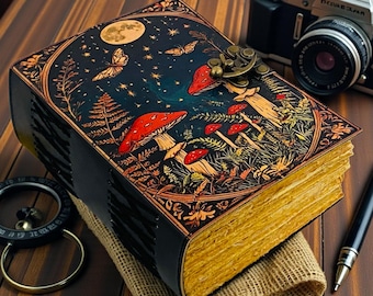 Puede incluir: Un diario de estilo vintage con una detallada ilustración de setas, mariposas y un cielo nocturno iluminado por la luna en la portada. El diario tiene un cierre y páginas de aspecto envejecido. También se ve una cámara y un bolígrafo.